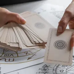 Dłonie trzymają talię kart, jedna karta z symetrycznym wzorem jest wyciągnięta. W tle widać astrologiczny krąg, jakby do wróżby z kart **high priestess tarot**.