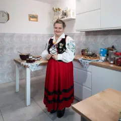 Kobieta w stroju ludowym stoi w kuchni obok stołu z dżemami. Na ścianie wisi zegar, wskazujący godzinę 10.
