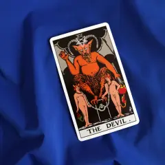 Karta Tarota "Diabeł" na niebieskim tle. Widnieje na niej demon z dwoma ludźmi u jego stóp, symbolizującymi pokusy i przywiązanie.