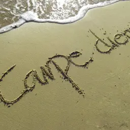 Napis "Carpe diem" na piasku, przypominający o docenianiu chwili. Fale obmywają brzeg.