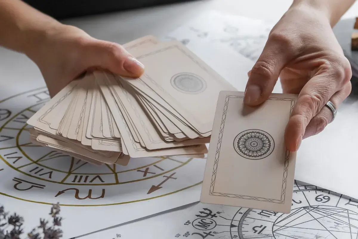 Dłonie trzymają talię kart, jedna karta z symetrycznym wzorem jest wyciągnięta. W tle widać astrologiczny krąg, jakby do wróżby z kart **high priestess tarot**.