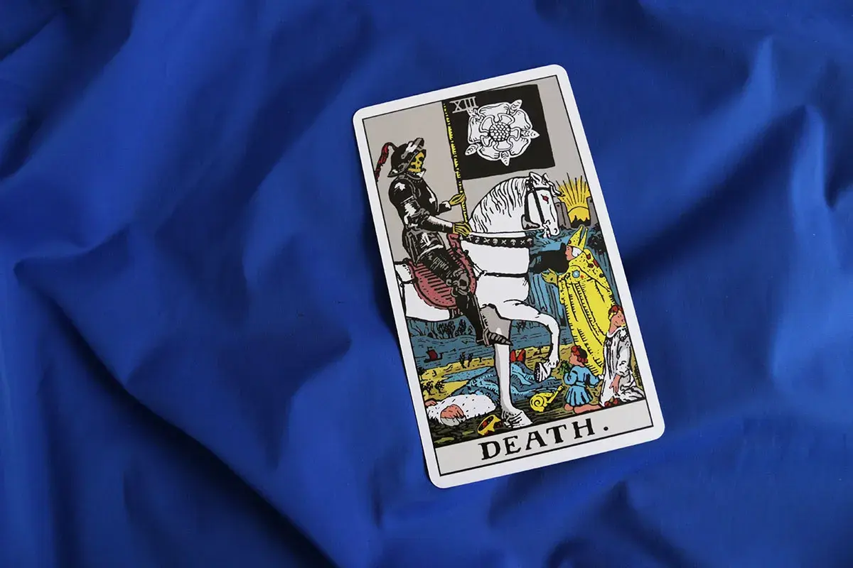 Tarot karta Śmierci na niebieskim tle. Postać w zbroi na białym koniu, niosąca flagę z różą, symbolizuje nieuchronne zmiany i transformację.