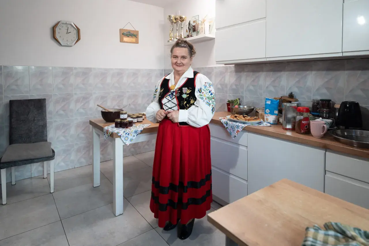 Kobieta w stroju ludowym stoi w kuchni obok stołu z dżemami. Na ścianie wisi zegar, wskazujący godzinę 10.