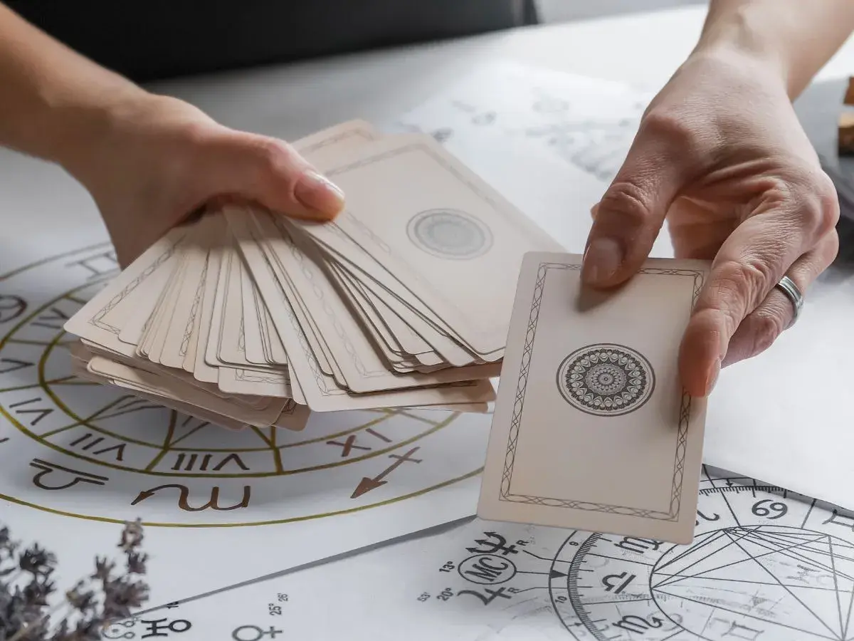 Dłonie trzymają talię kart, przygotowując się do wróżenia. Rozłożone na stole diagramy astrologiczne sugerują, jak stawiać tarota.