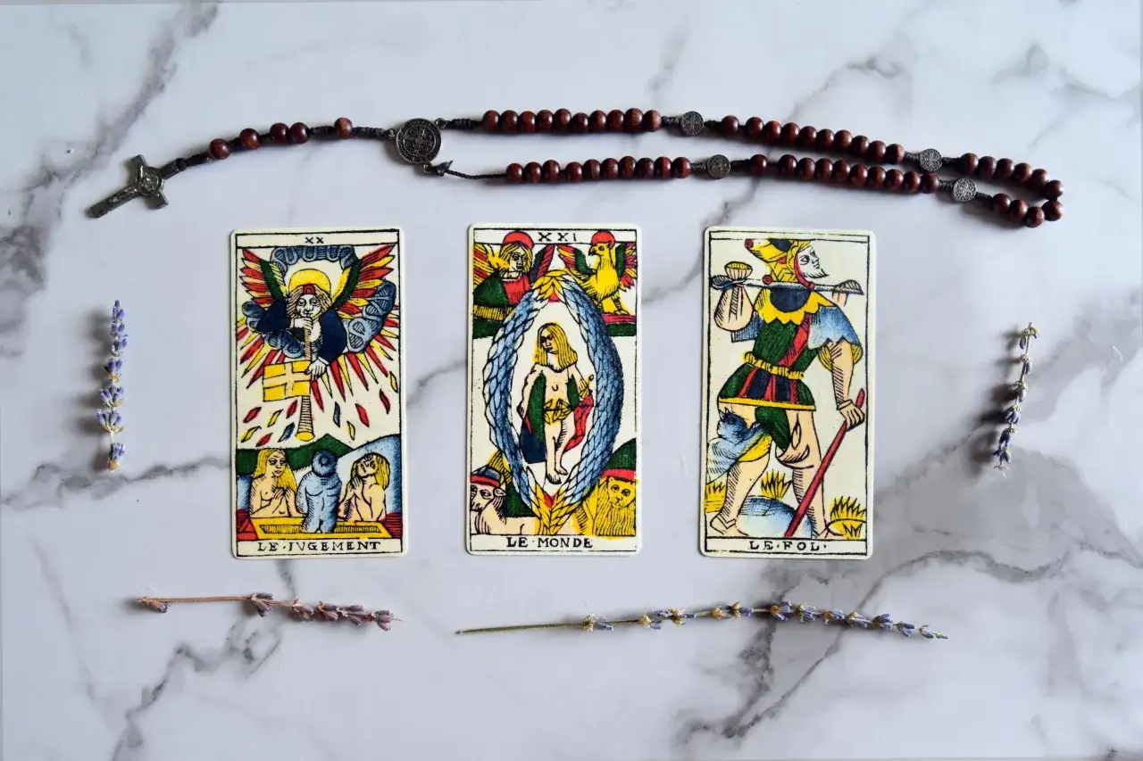 Tarot marsylski znaczenie kart: karty Sąd, Świat i Głupiec ułożone na marmurowym tle, obok różańca i lawendy.