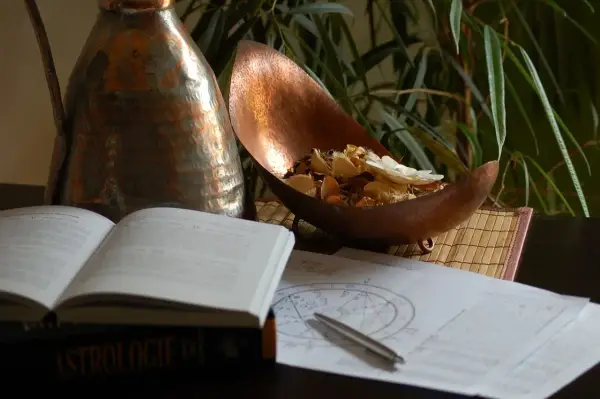 Na stole leży otwarta księga "Astrologie", obok wykresy, długopis i miedziana misa z suszonymi kwiatami. Umiarkowanie tarot uczucia.